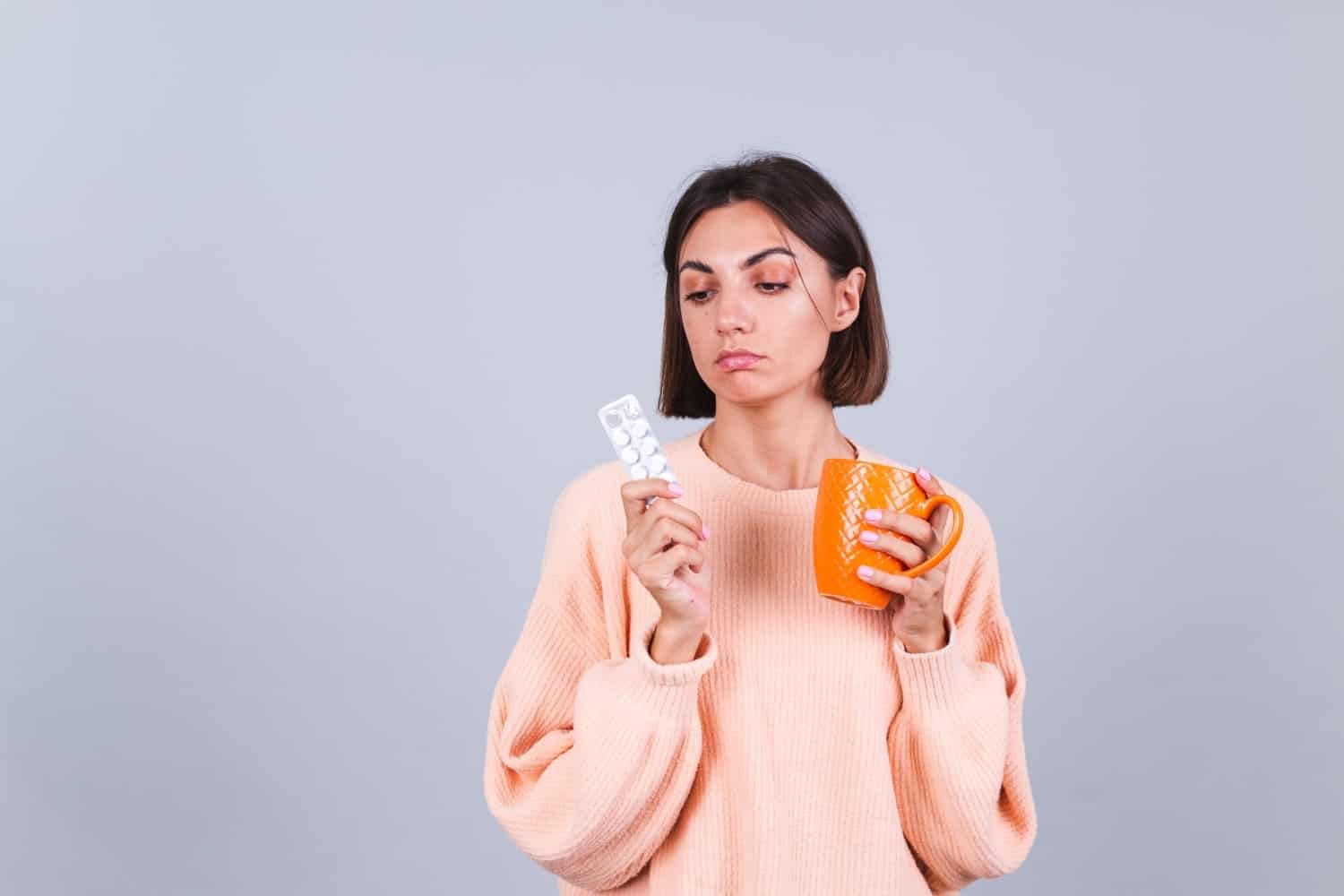 woman sweater gray wall holds mug pills with unhappy sad expression face redimensionada